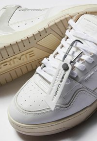 Scarpe sportive bianche in pelle con punta perforata, dettagli testurizzati beige e una striscia in suede grigio. Presenta un'etichetta metallica fissata ai lacci.