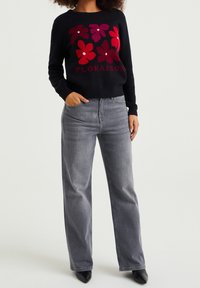 Schwarzer Pullover mit floralem Design in Rot und Pink, mit dem Wort "FLORAISON". Kombiniert mit hellgrauen, weit geschnittenen Jeans und schwarzen, spitzen Schuhen.