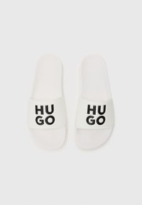 Witte rubberen slidesandalen met een brede band met de tekst "HUGO" in vette zwarte letters, een gestructureerde voetbed en een platte zool.