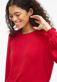 Camisa de manga larga texturizada roja con cuello redondo y puños fruncidos, que presenta arrugas sutiles y un ajuste relajado.