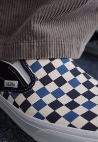 Zapatillas Vans slip-on con un patrón de cuadros azul y blanco, material de lona y suela de goma blanca, parcialmente cubiertas por unos pantalones de pana.