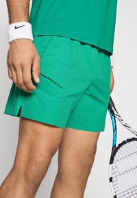 Πράσινα αθλητικά σορτς με υφή και γεωμετρικό σχέδιο, πλαϊνές τσέπες και σπασμένο στρίφωμα, συνδυασμένα με λευκό βραχιόλι και ρακέτα τένις.