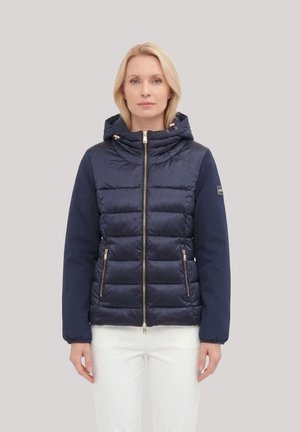 Donna con giacca trapuntata blu navy con cappuccio, chiusura con zip e tasche con zip, abbinata a pantaloni bianchi, in piedi davanti a uno sfondo neutro.