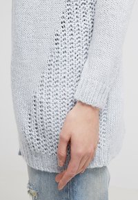 Jasnoszary, ażurowy sweter z luźnym splotem na górze, ściągaczami u rękawów i miękką teksturą; połączony z przetartymi dżinsami.