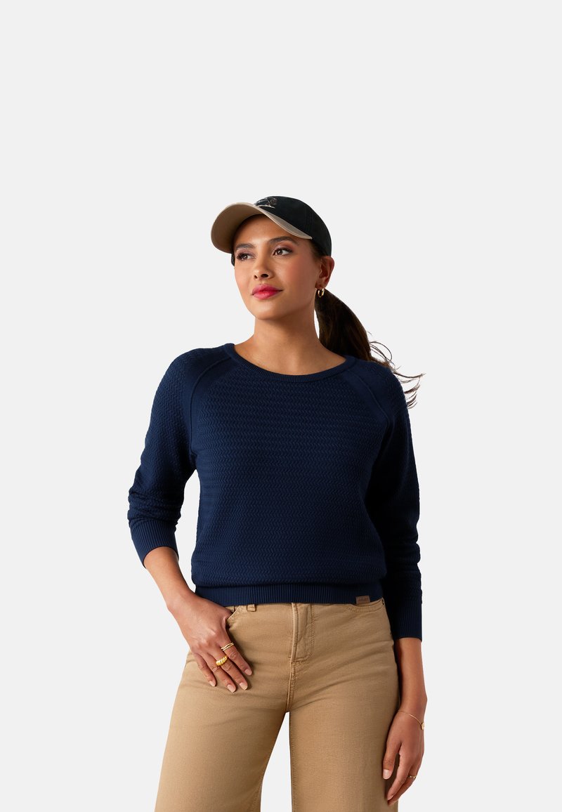 Marineblauer Strickpullover mit strukturiertem Muster, Rundhalsausschnitt und langen Ärmeln, kombiniert mit beigen Hosen und einer schwarz-beigen Mütze.