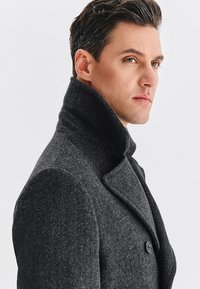 Szary płaszcz w jodełkę z wysokim kołnierzem, podwójnym zapięciem na guziki i gładką fakturą, noszony przez modelkę z krótkimi, stylizowanymi włosami na białym tle.