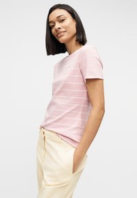 Νεαρή γυναίκα με ίσια μαλλιά, φορώντας ροζ και λευκή ριγέ μπλούζα και ανοιχτό μπεζ παντελόνι, με τα χέρια στις τσέπες, κοιτάζει την κάμερα.