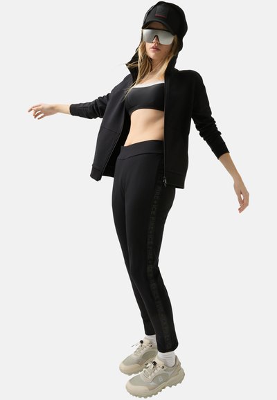 Schwarzes Sportoutfit bestehend aus einer Reißverschluss-Jacke und enganliegenden Leggings mit seitlichem Logomuster, kombiniert mit beigen Sneakers.