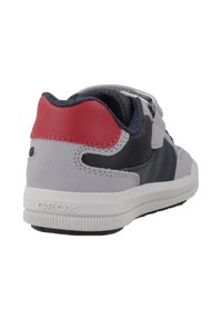 Zapatilla gris, azul marino y roja con suela blanca, correa de velcro y cuello acolchado vista desde la parte trasera y en un ángulo lateral.