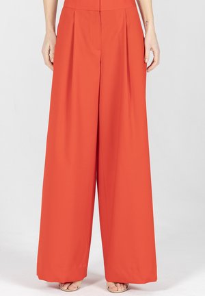 Pomarańczowe spodnie o szerokich nogawkach z wysokim stanem i przednimi zakładkami, noszone przez stojącą osobę, widoczne od talii do stóp, w otwartych sandałach.