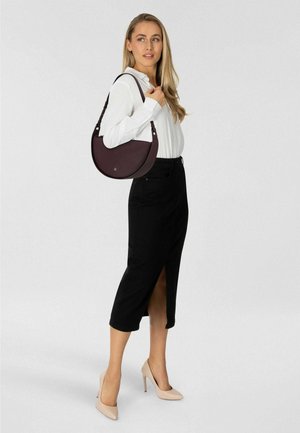 Bordeauxfarbene Halbmond-Handtasche aus glattem Material mit verstellbarem Riemen, über der Schulter getragen. Model in schwarzem Rock und Nude-Pumps.