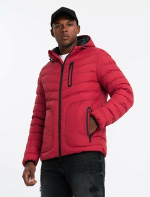 Rote Pufferjacke mit Kapuze, Reißverschluss vorne und aufgesetzten Taschen. Mit Steppnähten und schwarzen Akzenten. Kombiniert mit ausgebleichten schwarzen Jeans.