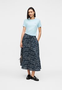 Γυναίκα που φοράει ανοιχτό μπλε μπλουζάκι και midi φούστα σε ναυτικό μπλε με σχέδια, κρατώντας μια μαύρη τσάντα, στέκεται μπροστά σε λευκό φόντο.