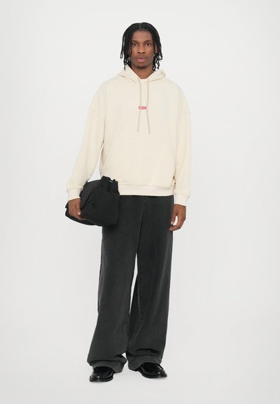 Light beige hoodie with pouch pocket and red lettering, paired with loose-fitting dark gray pants. Black bag held in left hand.