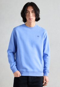Felpa azzurra con collo a giro, polsini e orlo a costine, con una piccola toppa con logo sul petto, abbinata a jeans scuri.