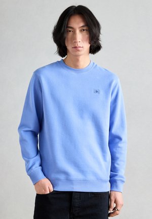 Lyseblå sweatshirt med rund halsudskæring, ribbede cuffs og kant, med et lille logo-patch på brystet, kombineret med mørke jeans.