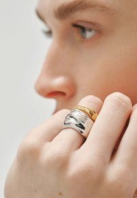 Tres anillos entrelazados en un dedo: uno de plata lisa, uno de plata texturizada y uno de oro pulido. Diseño minimalista con un brillo sutil.