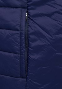 Mörkblå puffjacka med kviltat mönster, med en dragkedjeficka på vänster sida. Slät, blank yta.