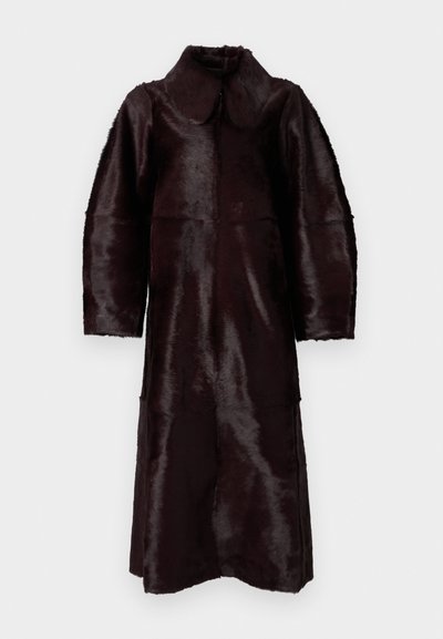 Tamsiai ruda ilga dirbtinio kailio paltas su plačiais atlapais ir ilgomis rankovėmis, pakabintas baltame fone.