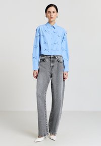 Frau steht und trägt ein hellblaues, kurz geschnittenes Hemd mit kleinen Schleifen, graue hoch taillierte Jeans und weiße, spitze Absätze vor einem einfarbigen Hintergrund.