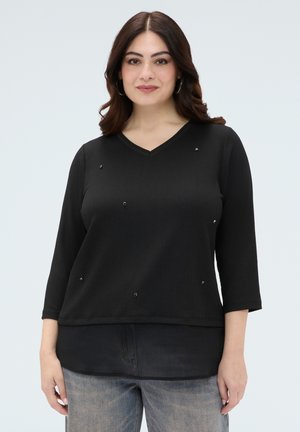 Vrouw met donker schouderlang haar draagt een zwart V-hals topje met kleine kralen en driekwart mouwen, gecombineerd met vervaagde blauwe spijkerbroek.