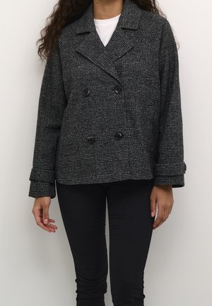 Femme portant une veste croisée à carreaux noirs et gris sur un haut blanc et un pantalon noir, debout devant un fond uni.