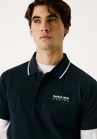 Marineblå polo-shirt med hvid piping, prydet med Red Bull Racing-logo. Korte ærmer, lavet af glat stof, egnet til afslappet brug.