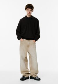 Schwarzes langärmliges Poloshirt, hellbeige lockere Jeans und schwarze Sneakers. Lockige Haare, Hände in den Taschen, neutraler Hintergrund.