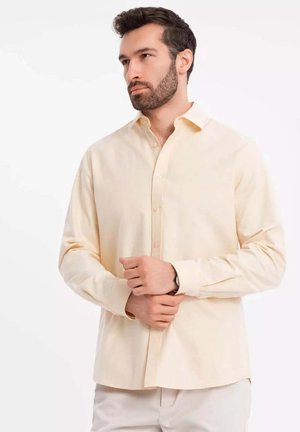 Camisa button-up de color beige claro, hecha de una tela suave, que presenta un cuello clásico y mangas largas, con sutiles detalles de costura.