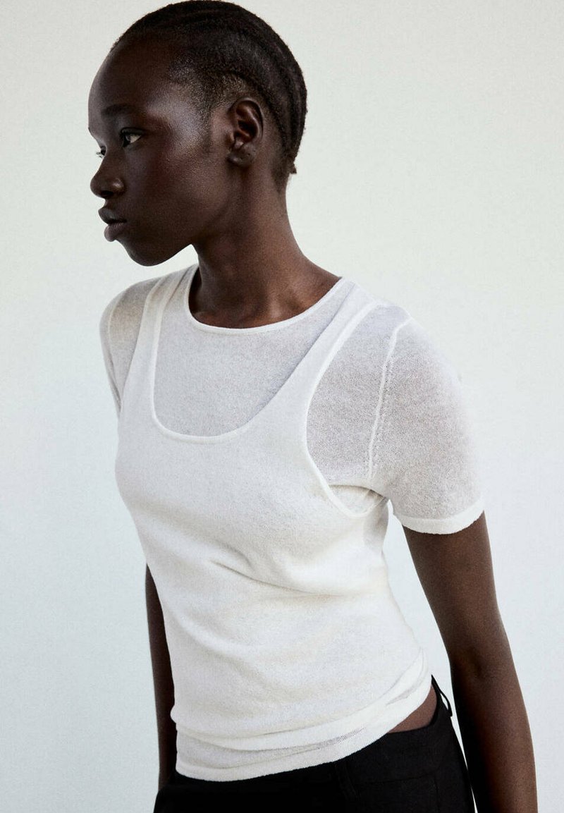 Jeune femme à la peau foncée et aux cheveux tressés porte un haut blanc à manches courtes sous un débardeur blanc, tournée vers la gauche sur un fond uni.