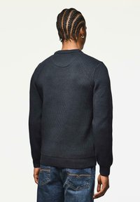 Person mit geflochtenem Haar, der einen schwarzen Rippensweater und blaue Jeans trägt, von hinten vor einem einfarbigen Hintergrund gezeigt.