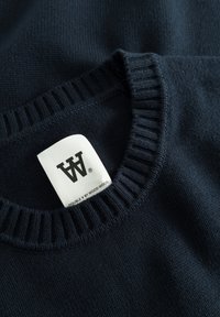 Pull en tricot bleu marine avec col et poignets côtelés, présentant une étiquette blanche avec un texte de marque noir à l'intérieur de l'encolure.