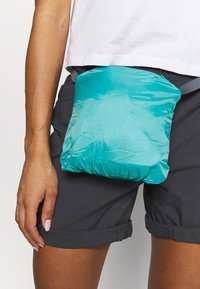 Personne portant un short foncé et une chemise blanche avec un petit sac turquoise attaché à la taille par une sangle grise.