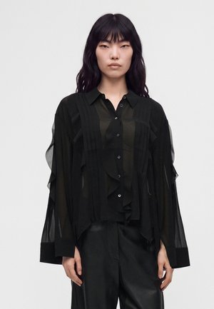 Femme aux longs cheveux noirs portant un chemisier noir transparent à volants et un pantalon noir, debout devant un fond blanc uni.