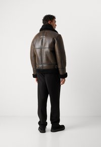 Giacca di pelle marrone con colletto in pelliccia sintetica nera, caratterizzata da dettagli trapuntati e vita con cintura. Indossata con pantaloni neri e scarpe scure.