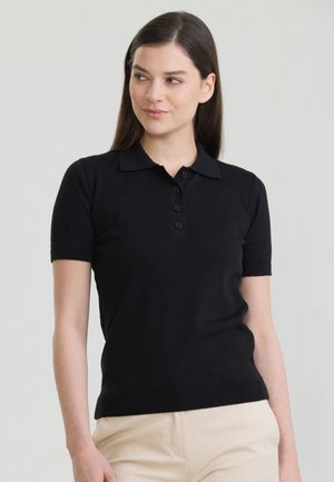 Jonge vrouw met lang donker haar, gekleed in een zwart polohemd met korte mouwen en beige broek, kijkt iets naar links.