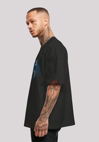 Svart oversized T-skjorte laget av bomull, med et blått grafisk design. Modellen har tatoveringer på armene og har kort hår.