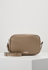 Hnědá texturovaná kožená crossbody taška obdélníkového tvaru se stříbrnými detaily a logem "HUGO" na přední straně. Součástí je nastavitelné ramenní popruh.