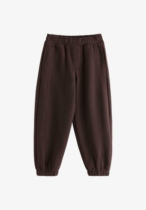 Smilende pige med langt hår iført en blå og brun stribet langærmet trøje, mørkebrune bukser og hvide sneakers, stående mod en hvid baggrund.