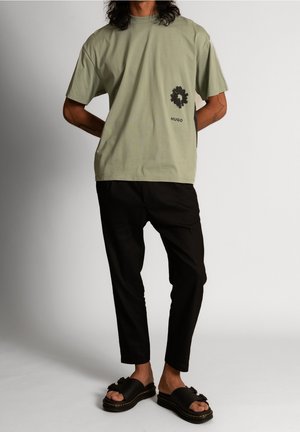 Mann trägt lockeres olivgrünes T-Shirt mit schwarzem Blumenmuster und dem Schriftzug "HUGO", schwarze kurze Hose und schwarze Sandalen vor weißem Hintergrund.