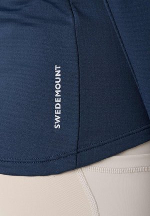 Zbliżenie na granatowy sportowy top z logo „Swedemount” w kolorze białym oraz beżowe legginsy z widocznymi przeszyciami.