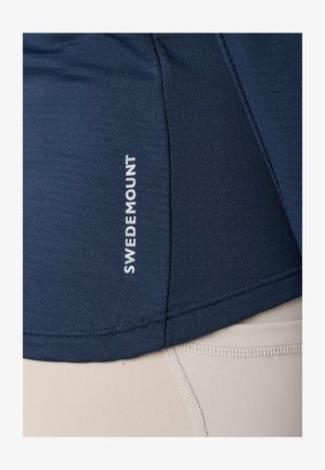 Zbliżenie na granatowy sportowy top z logo „Swedemount” w kolorze białym oraz beżowe legginsy z widocznymi przeszyciami.