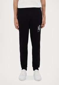 Schwarze Sweatpants mit schmaler Passform, elastischem Bund, Seitentaschen und einem weißen Logo-Druck auf dem linken Oberschenkel. Kombiniert mit weißen Sneakers.