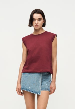 Femme aux cheveux courts et foncés portant un haut bordeaux sans manches et une jupe en jean asymétrique ornée de petites perles, debout devant un fond uni.