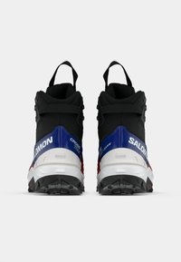 O pereche de ghete de drumeție Salomon Cross Track, negre, albastre, albe și roșii, prezentate din spate, cu bucle pentru călcat și tălpi robuste, pe un fundal alb.