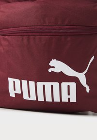 Kastanjett sportsbag laget av slitesterk stoff, med en hvit Puma-logo og glidelås. Teksturert overflate med fremtredende sømdetaljer.