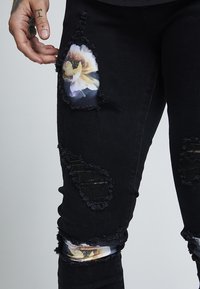 Black distressed jeans featuring large rips with transparent floral patterns. Raw hems with frayed edges; close-fitting design.