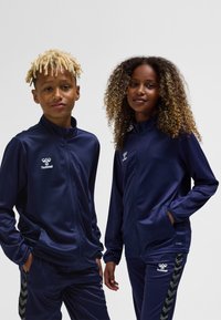 Navy blue tracksuits for children with zip-up jackets and pants. Features logo on chest and contrasting chevron pattern on pant sides.