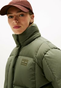 Doudoune vert olive avec col montant, arborant une patch logo sur la poitrine gauche. La personne porte une casquette en velours côtelé marron.
