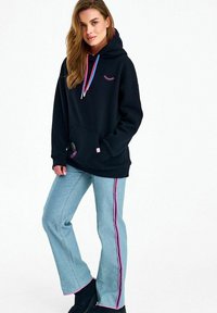 Žena v černé mikině s růžovými a modrými šňůrkami, doplněná světle modrými džínami s růžovými a černými proužky na bocích a černými botami.